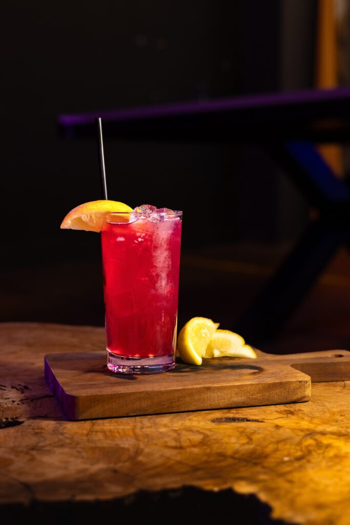 A tall glass of red iced drink with a straw and an orange wedge garnish, placed on a wooden board with lemon wedges beside it, set on a rustic wooden table.
