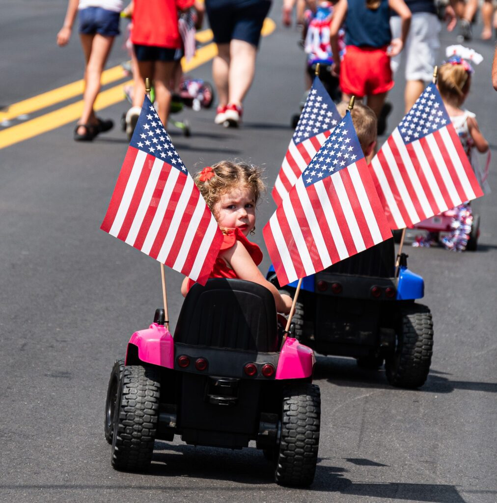 A young girl in a toy car holds two American flags during a parade, surrounded by other children and adults on a sunny street.