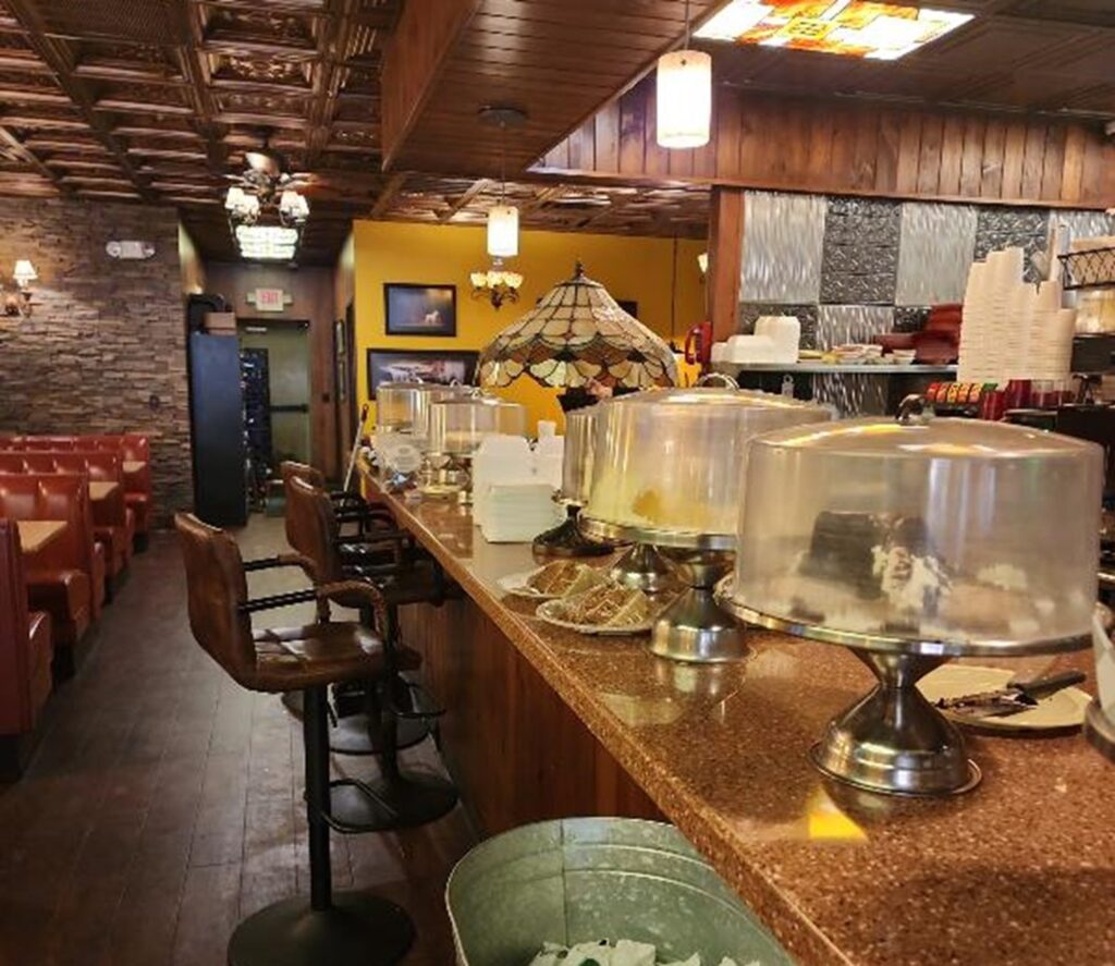 A cozy café with brown leather booths, a long counter with covered cakes and pies, bar stools, and warm lighting. The interior features wood and stone accents, creating a inviting atmosphere.