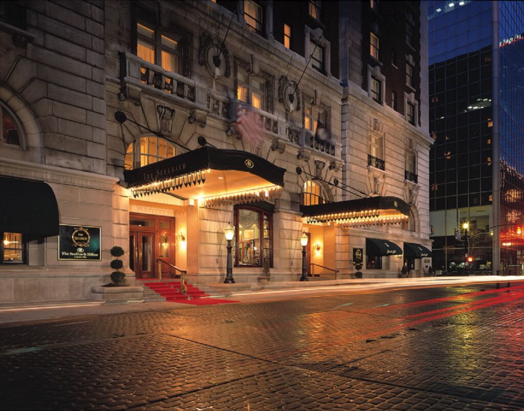 Historic stone hotel exterior at dusk with warm lights, a red carpet on the steps, black awnings, and elegant balconies. The wet cobblestone street reflects the glow from the entrance.
