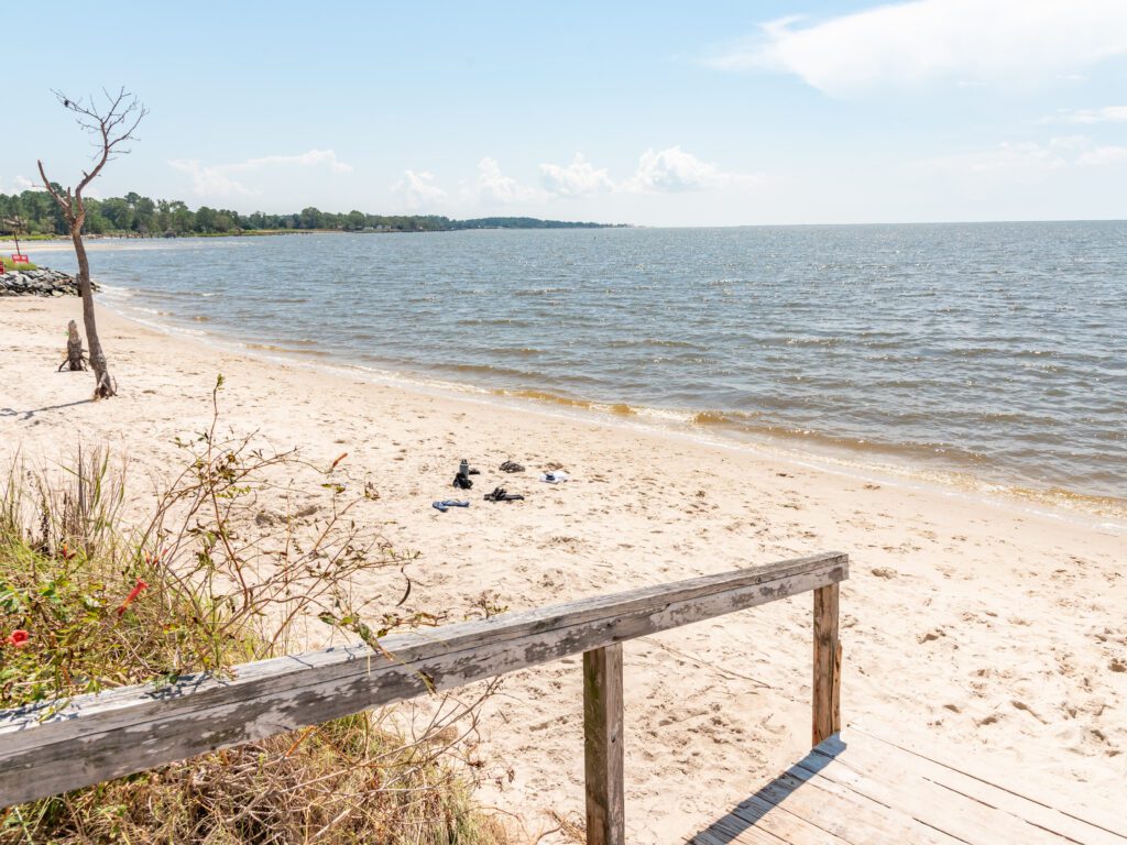 A sandy beach with gentle waves, a wooden handrail leading to the shore, scattered vegetation, and a lone tree on the left. The sky is partly cloudy, and the coastline extends into the distance.
