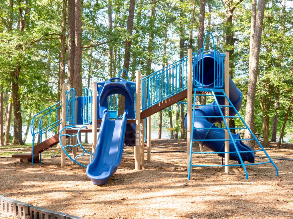 A blue and tan playground structure with slides, climbing bars, and a tunnel slide, set in a wooded park area with trees and wood chip ground covering.
