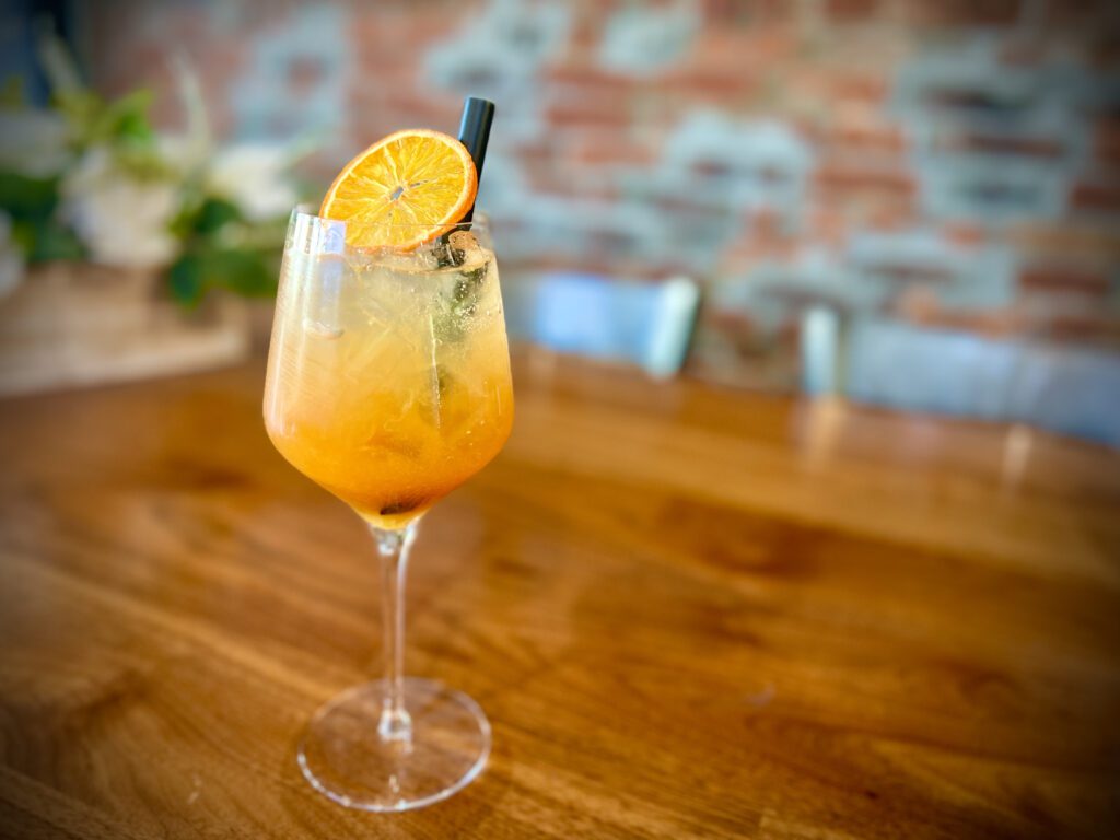 A cocktail in a wine glass garnished with a slice of dried orange and a black straw, set on a wooden table with a blurred brick wall and plants in the background.