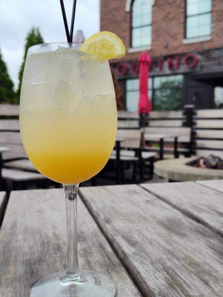 A chilled yellow cocktail with ice and a lemon slice garnish sits on a wooden table outdoors, with patio seating and a brick building in the blurry background.