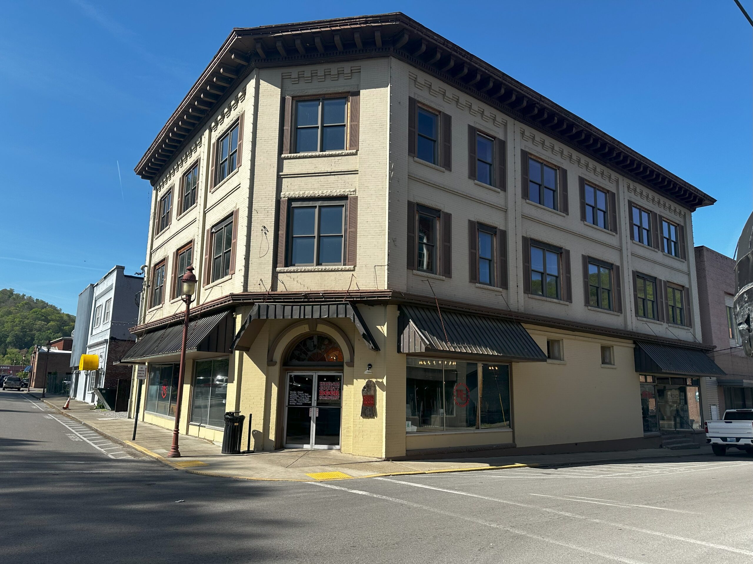 A three-story cream-colored historic building with large windows sits on a quiet street corner under a clear blue sky. The sidewalk is empty, and sunlight casts shadows along the building’s facade.