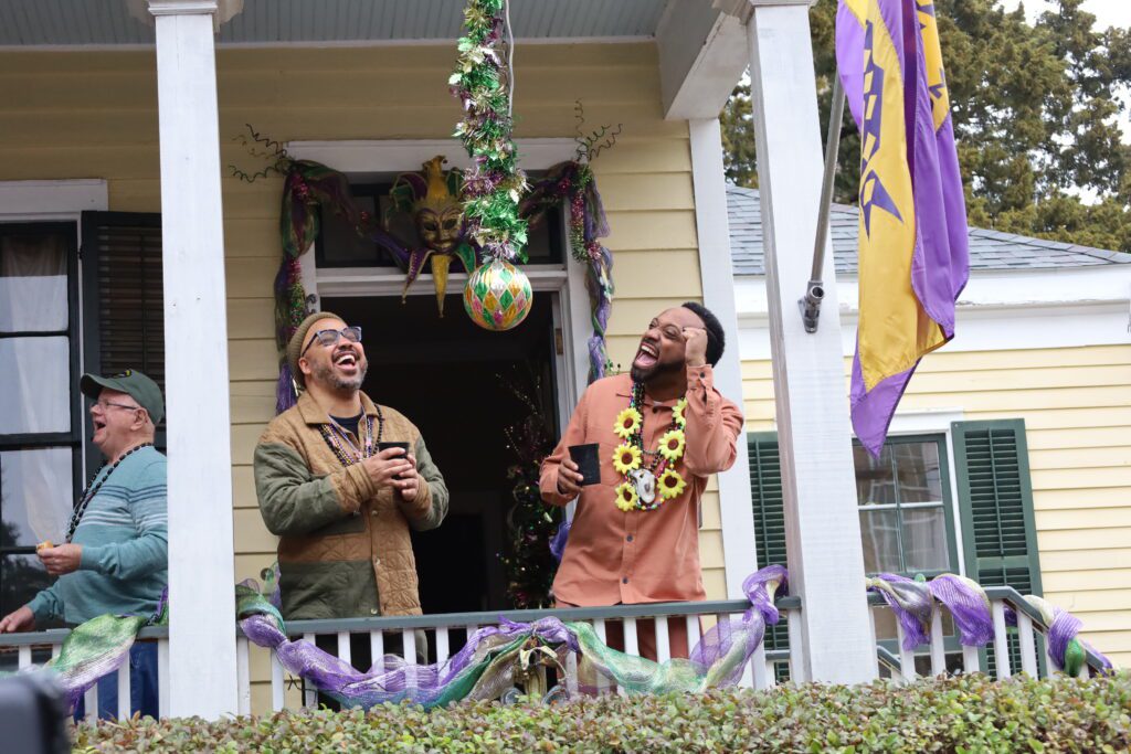 Three people on a festive porch decorated with Mardi Gras beads, ribbons, and masks. Two men in front are laughing, wearing colorful shirts and beads. A Mardi Gras flag and a ball hang overhead.