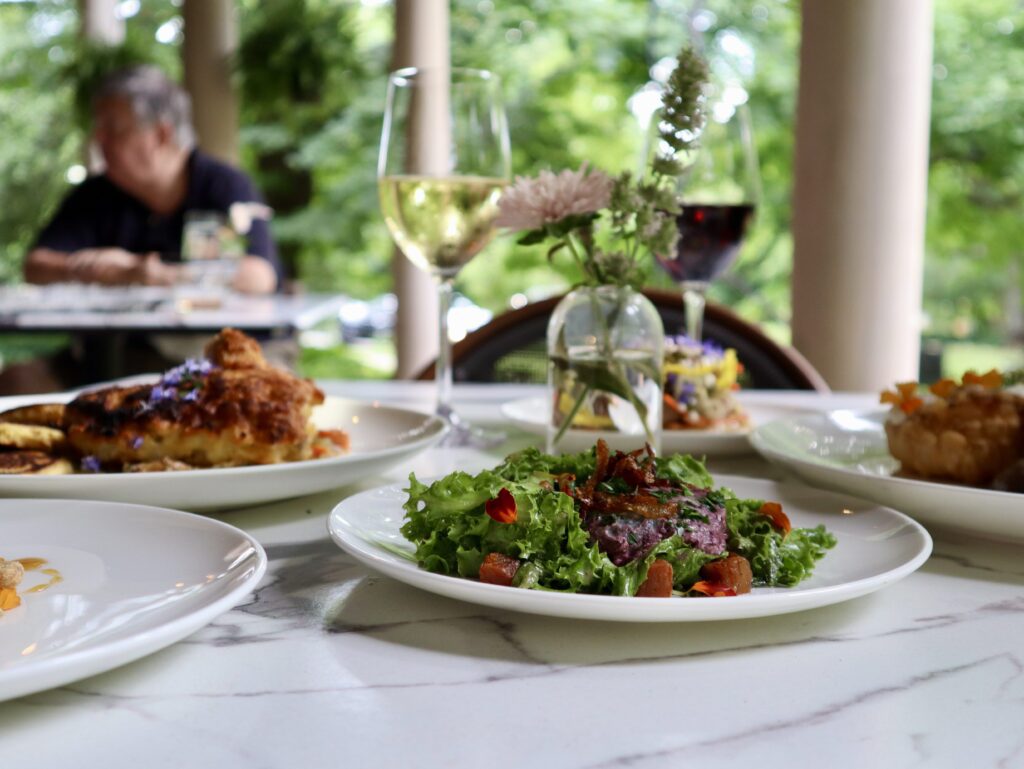A close-up of a white marble table set with plates of salad, roasted vegetables, and glasses of white and red wine. A small flower vase decorates the table, with a blurred person sitting in the background outdoors.