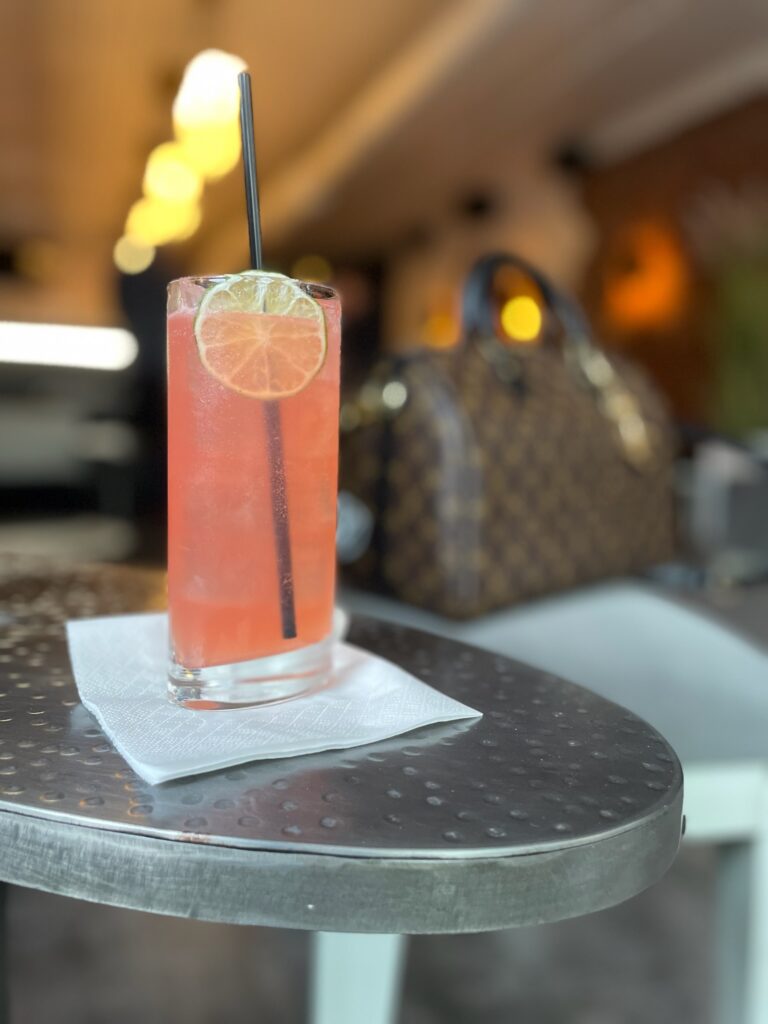 A tall glass of pink cocktail with ice, a lime slice, and a black straw sits on a napkin on a round metal table. A blurred handbag and indoor seating are visible in the background.