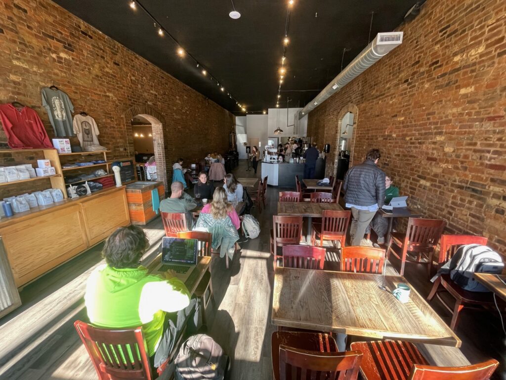 Customers sit at wooden tables in a cozy coffee shop with brick walls, working on laptops or chatting. The café is busy, with people waiting near the counter. Shelves display coffee bags and merchandise.