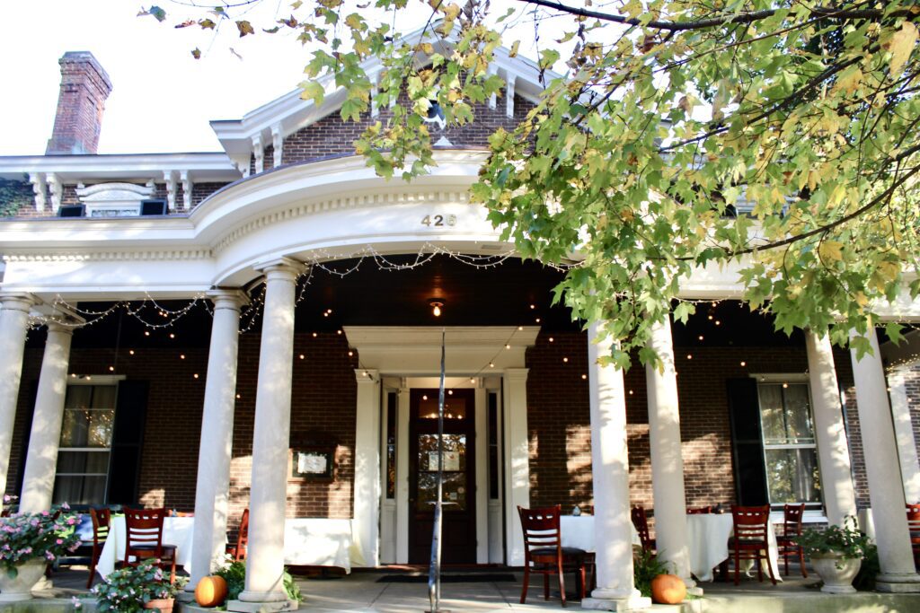 A brick building with white columns, a porch set with tables and chairs, potted flowers, and pumpkins. String lights hang above the entrance, and leafy green trees frame the top of the image.