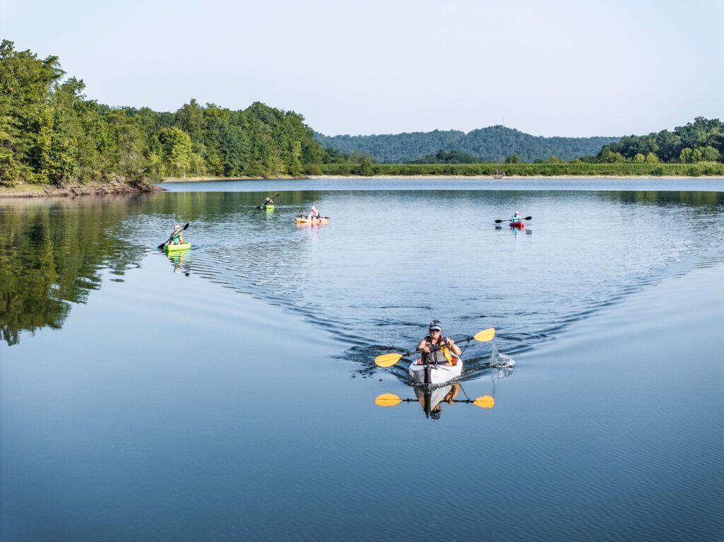 Several people are kayaking on a calm lake surrounded by trees and distant hills, with clear blue sky and reflections on the water.