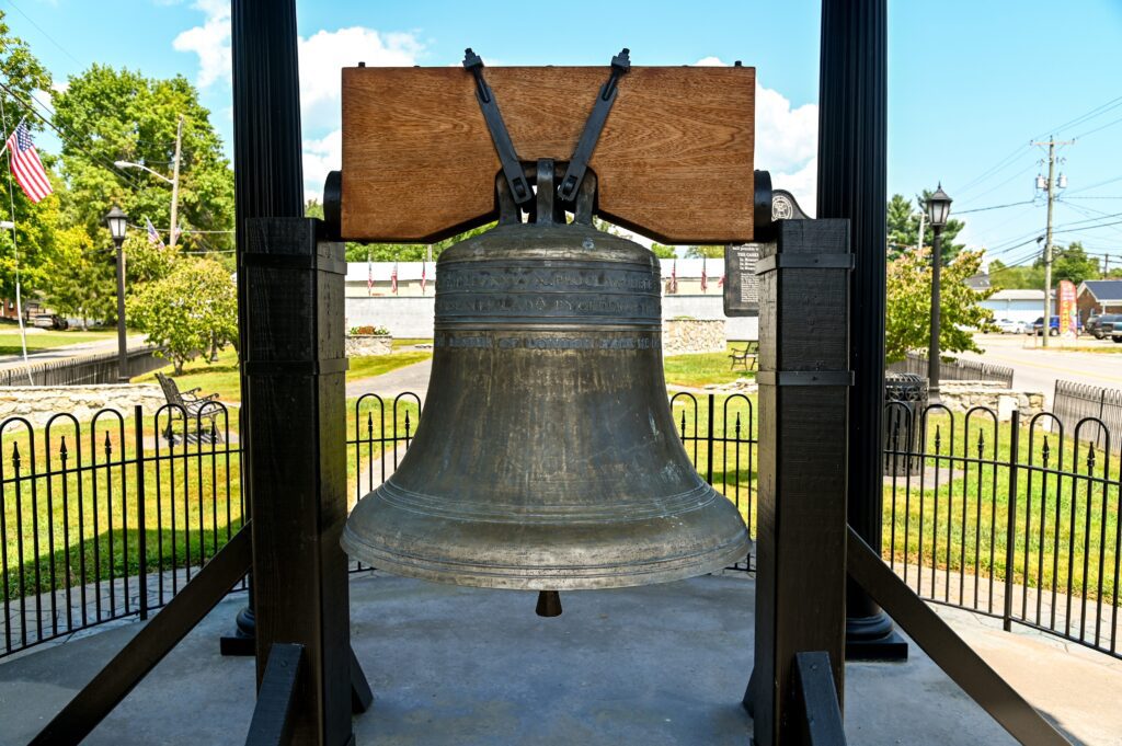 A large, weathered bronze bell hangs in a black metal frame outdoors, surrounded by a black fence, with green grass, trees, and an American flag visible in the background.