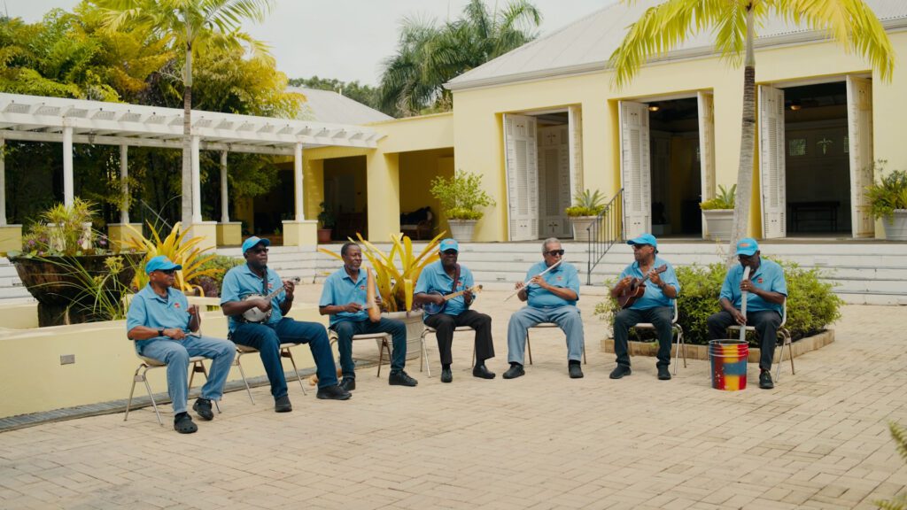 Seven men in blue shirts and hats sit in a row outside a yellow building, playing musical instruments, including guitars, a drum, and maracas. They perform in a sunny, tropical courtyard with palm trees and plants.
