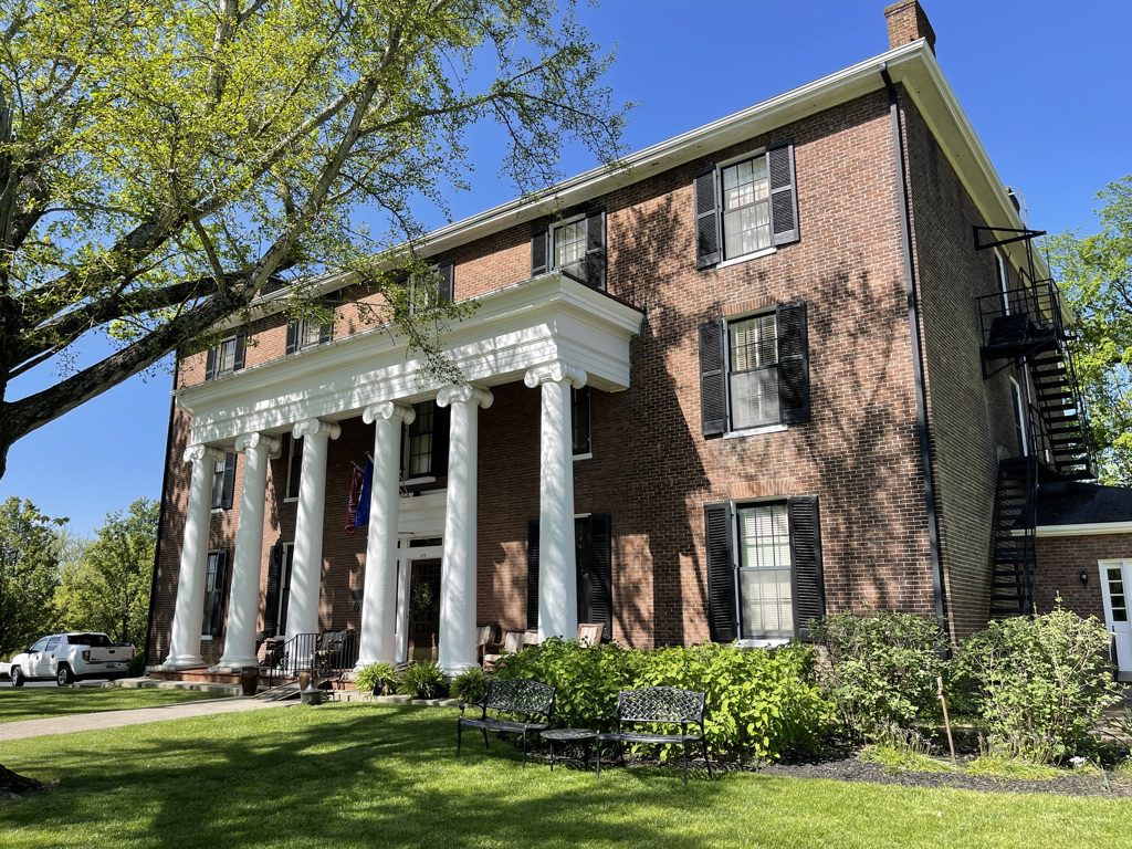 A large brick building with tall white columns at the entrance, black shutters on the windows, an American flag by the door, a black fire escape on the side, and green grass with benches and trees in front.