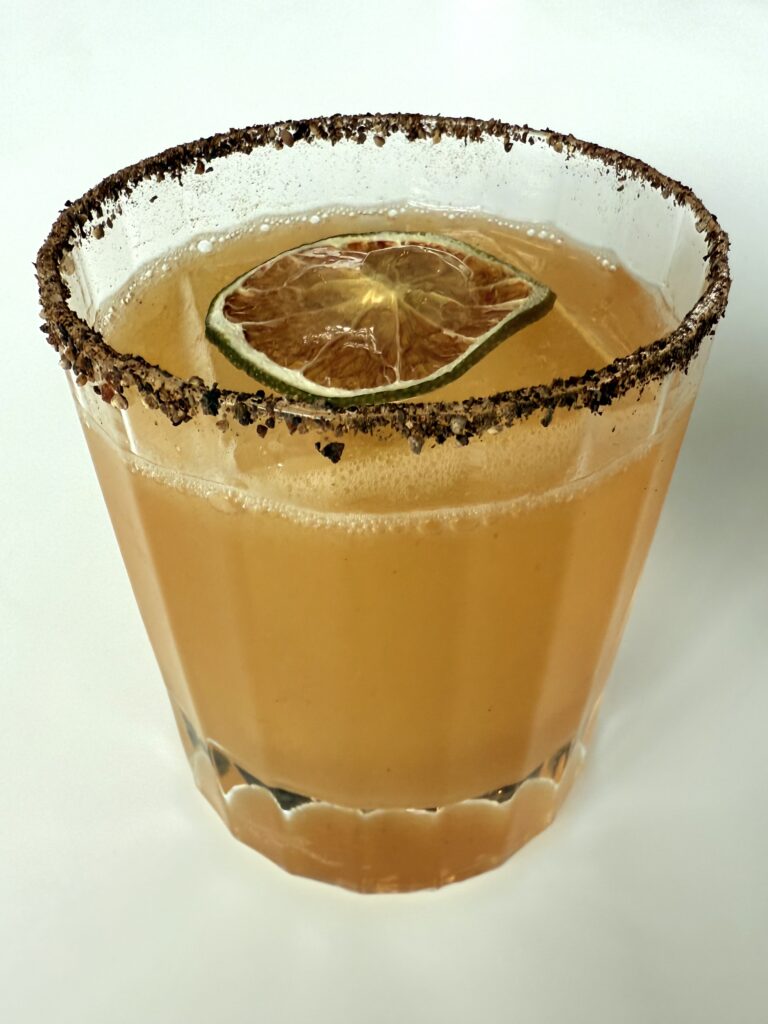 A glass of orange-colored cocktail with a dried lime slice floating on top. The rim of the glass is coated with a dark spice or salt mixture, and the drink is served on a plain white background.