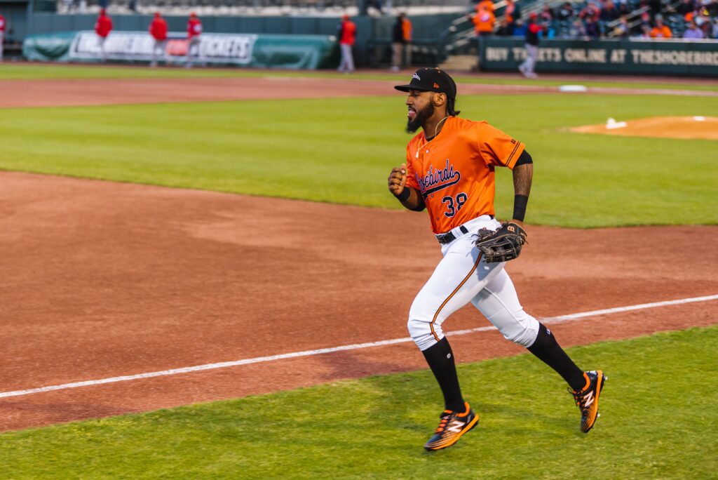 A baseball player in an orange and black uniform runs along the edge of the field near the infield dirt. Some players and spectators are visible in the background.