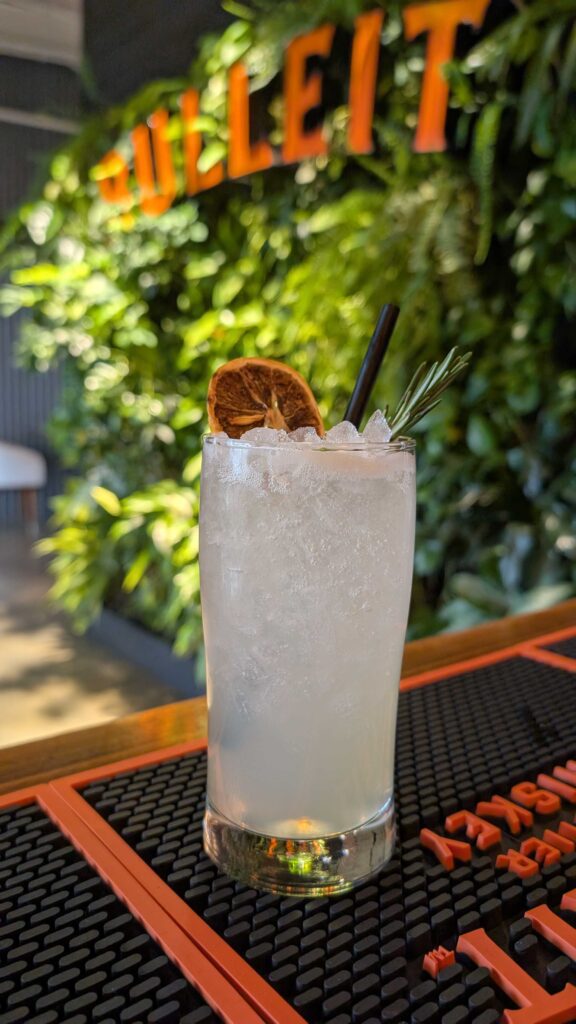 A cocktail with ice, garnished with a dried citrus slice and rosemary sprig, sits on a bar mat. In the background, green plants and a partial BULLEIT sign are visible.