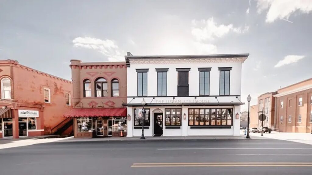 A wide street view of a modern white building labeled The Urban Stillhouse, flanked by two older brick buildings under a bright sky with scattered clouds.