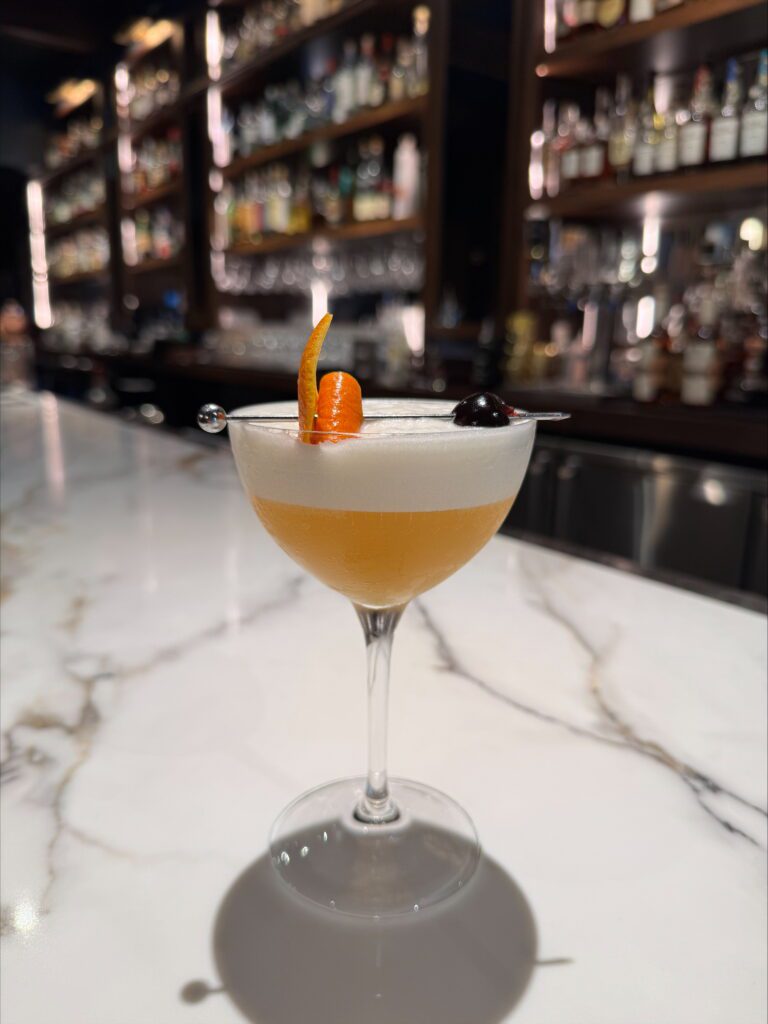 A cocktail with a frothy top, garnished with an orange twist and a cherry, sits on a marble bar counter. Shelves of liquor bottles are blurred in the background.