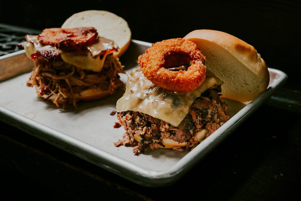 Two pulled pork sandwiches on a metal tray, each topped with melted cheese. One has crispy bacon; the other is topped with two golden onion rings. Both sandwiches have the top buns set slightly to the side.