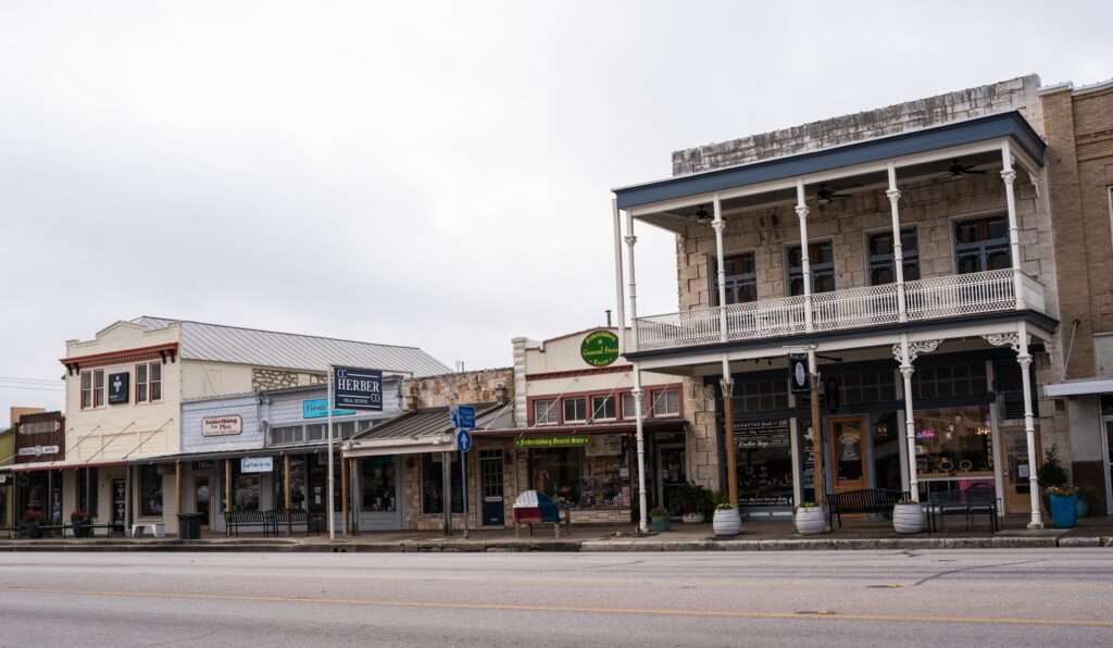 A row of small shops and businesses with vintage facades line a quiet street in a historic downtown area, featuring a two-story building with a balcony and overcast skies above.