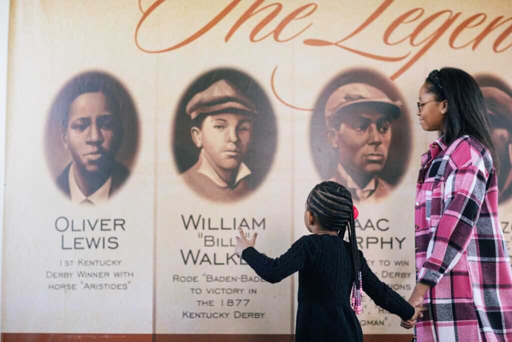 A woman and a child stand in front of a mural featuring portraits and names of famous Black jockeys, including Oliver Lewis, William Billy Walker, and Isaac Murphy, at a Kentucky Derby exhibit.
