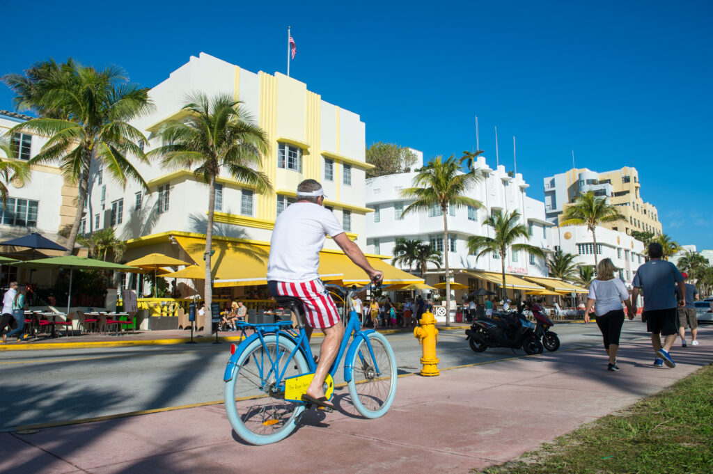 A man in red, white, and blue shorts rides a blue bicycle on a sunny street lined with palm trees and art deco buildings with yellow awnings, while people walk nearby.