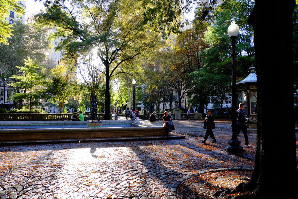 People walk and sit near a fountain in a sunlit park with trees, cobblestone paths, fallen leaves, and lampposts. Sunlight streams through the foliage, creating shadows on the ground.