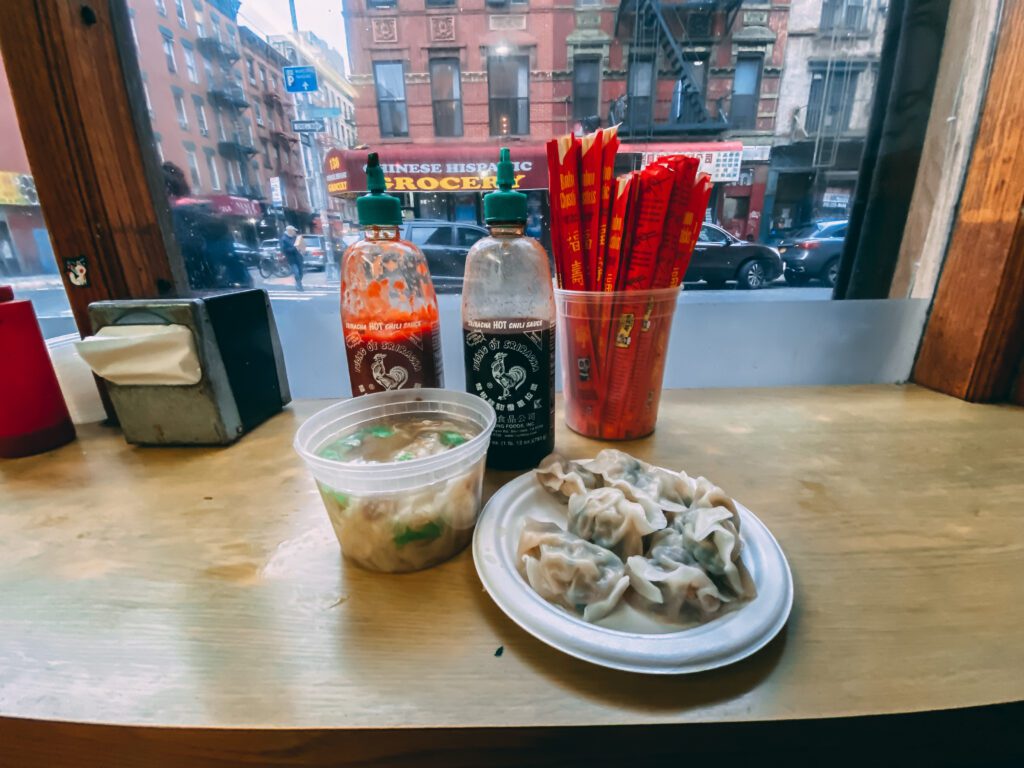 A plate of dumplings and a container of soup sit on a wooden counter by a window, with bottles of Sriracha and soy sauce, red chopsticks, and napkins; a city street is visible outside.