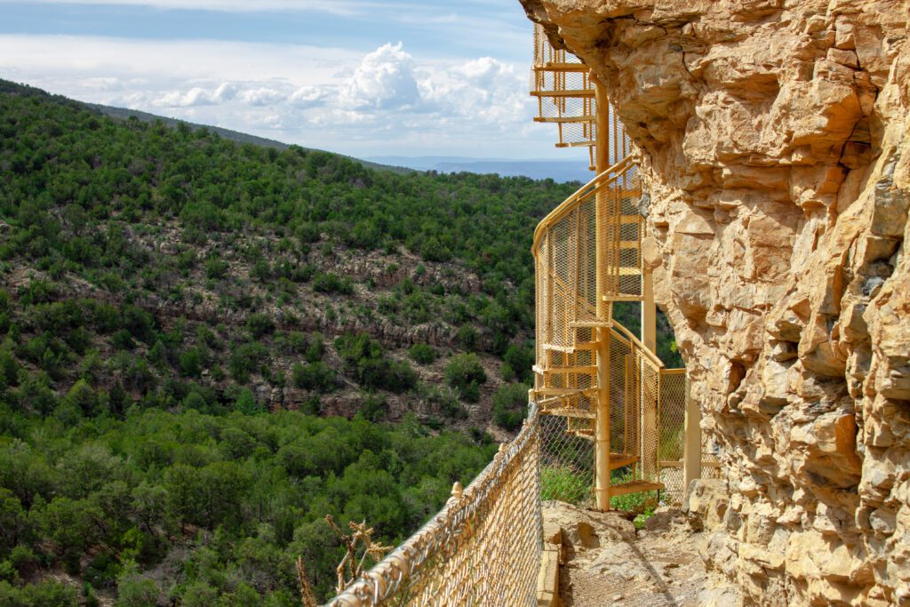 A metal spiral staircase is attached to a rocky cliff, overlooking a lush green forest and distant hills under a partly cloudy sky.