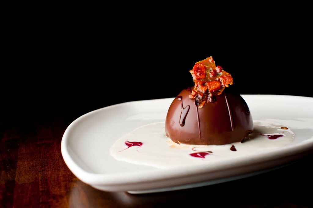 A chocolate dome dessert sits on a white plate, surrounded by white sauce with pink accents, and topped with a cluster of candied nuts or brittle, against a dark background.
