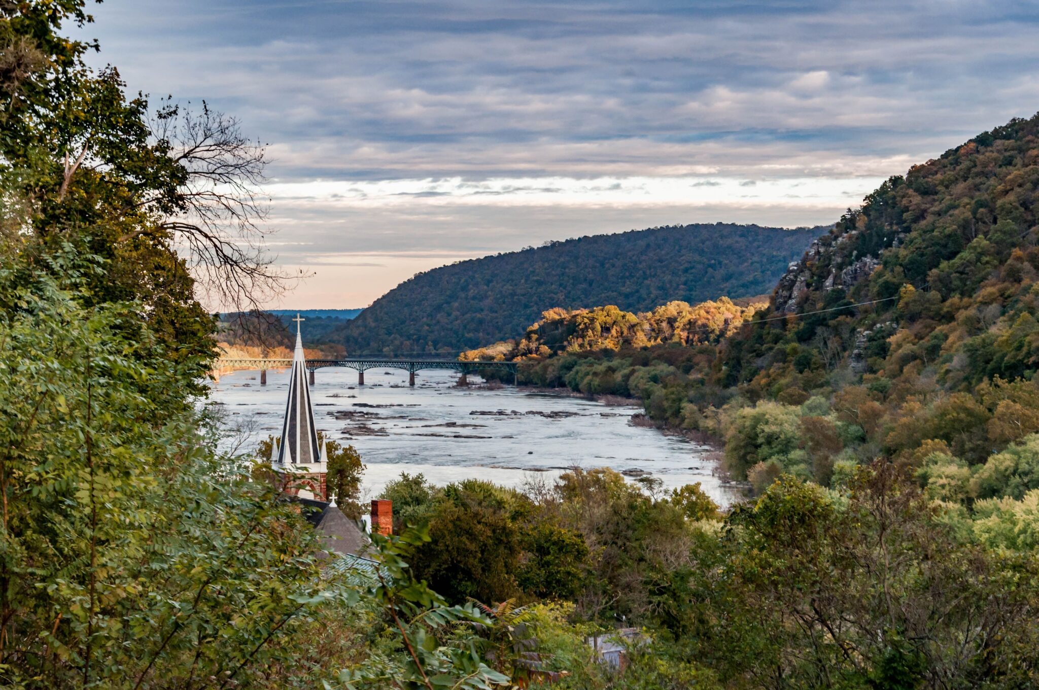 A scenic river meanders between tree-covered hills under a partly cloudy sky, with a church steeple and a distant bridge visible among lush green foliage in the foreground.