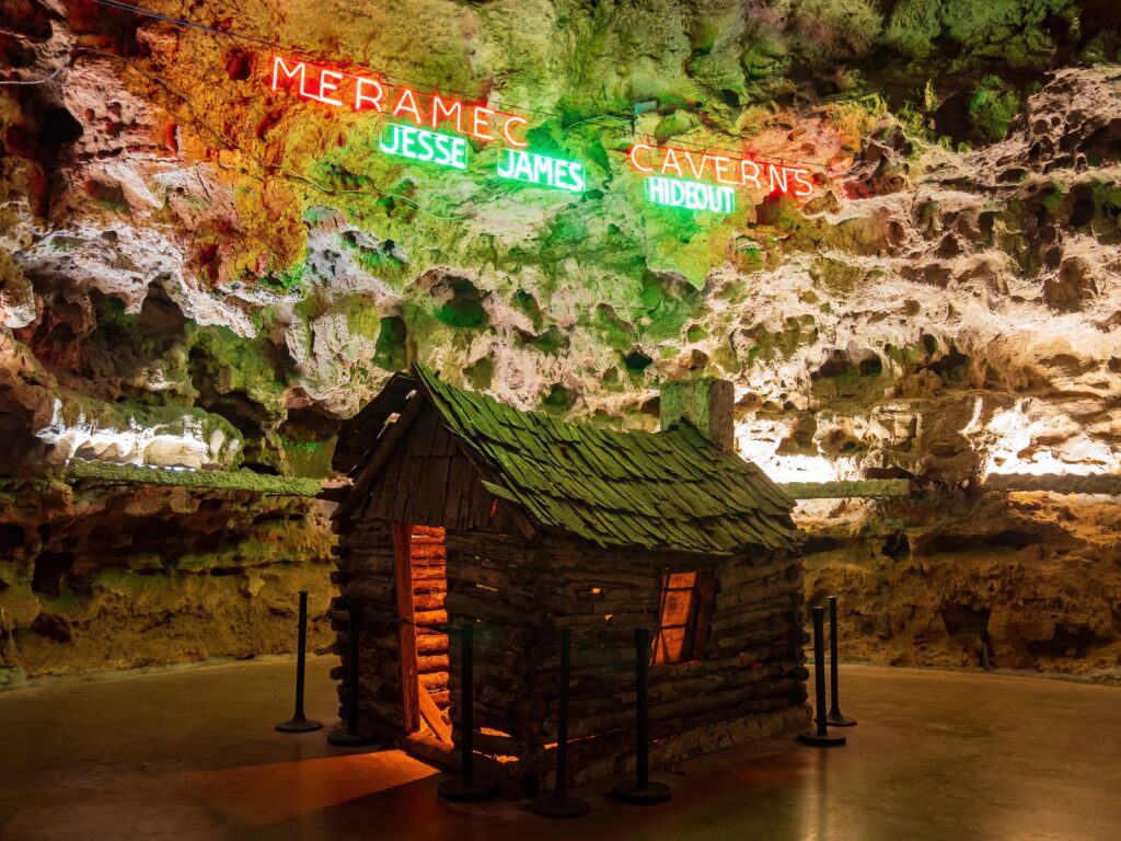 A small wooden log cabin is displayed inside a cave, surrounded by rope barriers. Neon signs above read Meramec Caverns and Jesse James Hideout on the rough, illuminated cave wall.