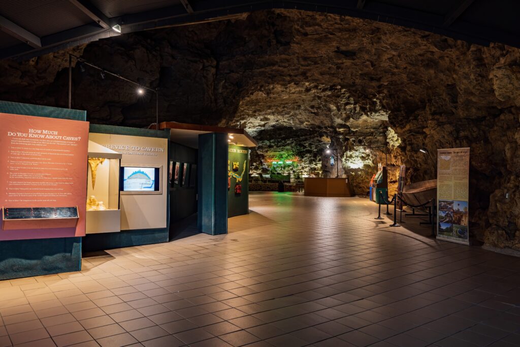 An indoor exhibit area inside a cave features informational displays and glass cases on tiled flooring, with rocky cave walls and ceiling in the background, and additional exhibits lit by spotlights.