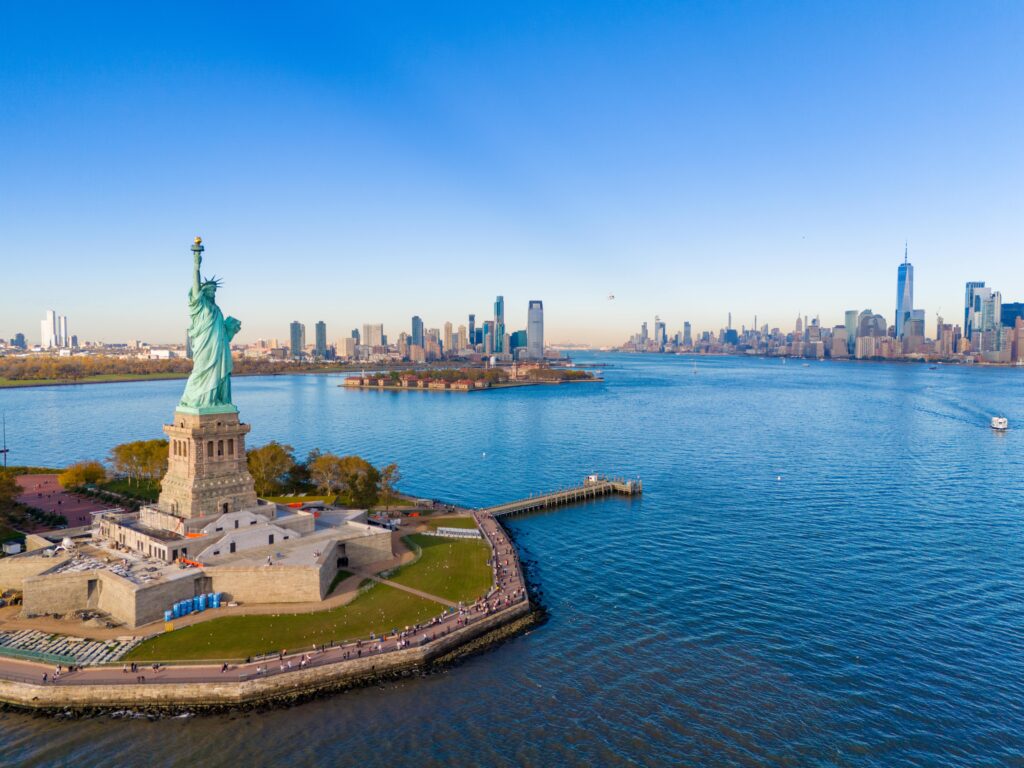 Aerial view of the Statue of Liberty on Liberty Island, with the Manhattan skyline and surrounding blue water under a clear sky in New York City.