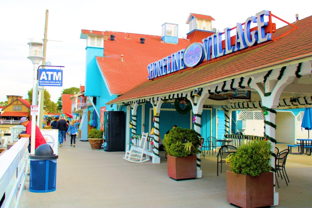 A colorful boardwalk at Shoreline Village with shops, rocking chairs, plants, an ATM sign, and people walking along the seaside on a bright day.