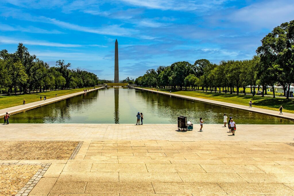 Wide view of the National Mall in Washington, D.C. showing the Reflecting Pool, people walking, and the Washington Monument in the distance under a blue sky with some clouds.