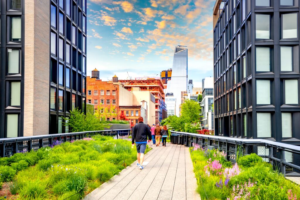 People walk along a raised urban park lined with green plants and flowers, surrounded by modern buildings under a colorful, partly cloudy sky.