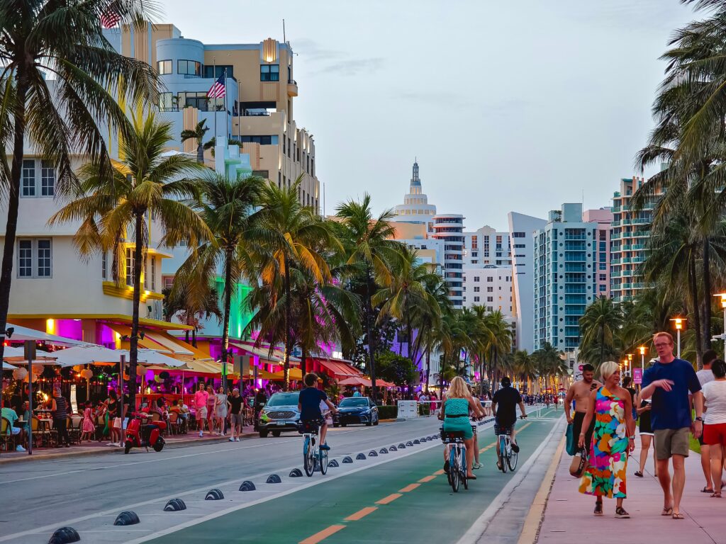 People walk and ride bicycles along a lively street lined with palm trees and colorful buildings, with cafes and restaurants glowing with neon lights at dusk.