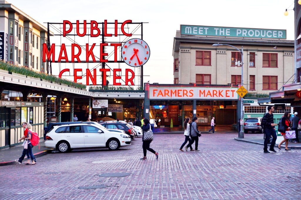 People cross a brick-paved street in front of Seattle’s iconic Pike Place Market, with large signs reading “PUBLIC MARKET CENTER” and “FARMERS MARKET” visible on surrounding buildings.