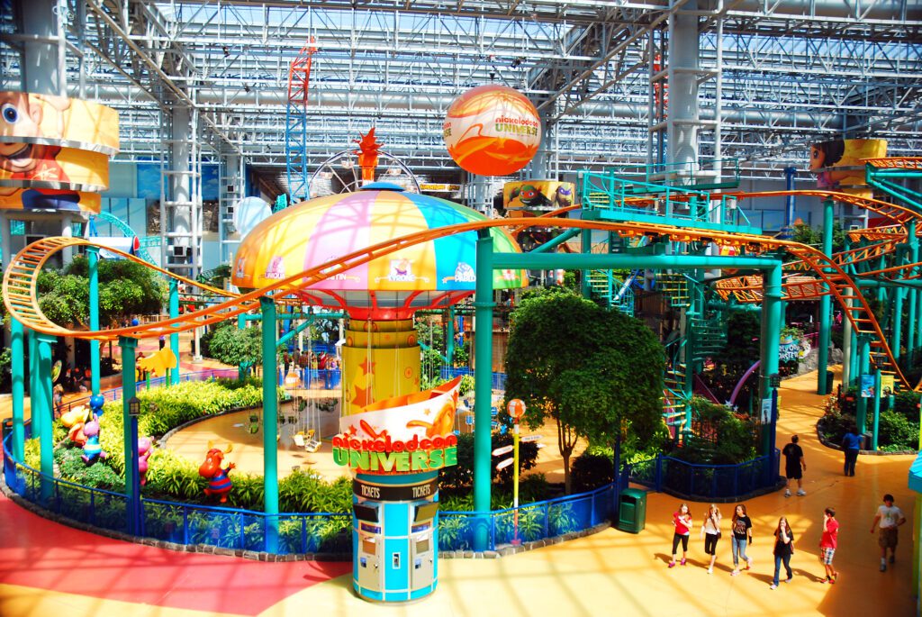 An indoor amusement park with bright orange roller coaster tracks, green support beams, colorful decorations, lush plants, and people walking below. A large sign reads “Nickelodeon Universe.”.
