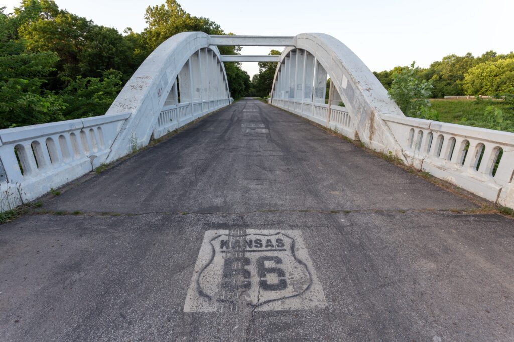 A historic white arched bridge with a Kansas Route 66 shield painted on the road, surrounded by green trees under a clear sky.