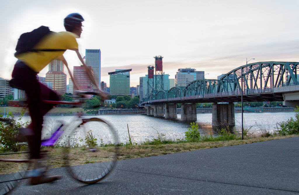 A person on a bicycle rides along a riverside path near a bridge, with a cityscape of tall buildings in the background at sunset.