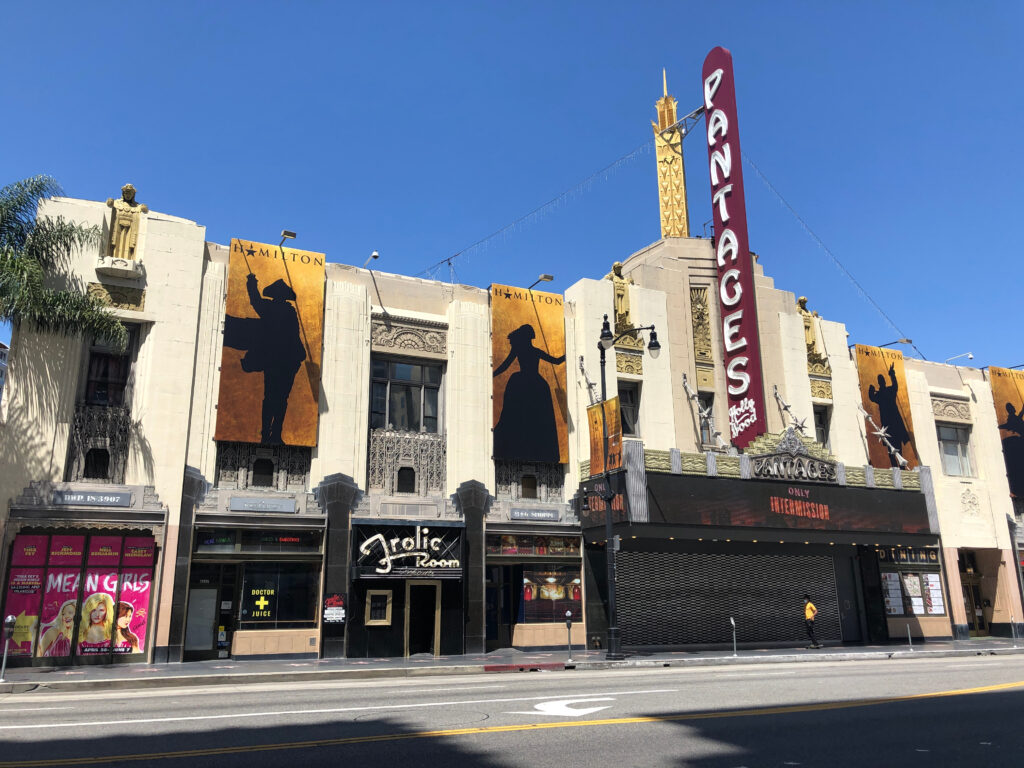 The Pantages Theatre in Los Angeles with Hamilton banners hanging on its facade, a large vertical marquee reading Pantages, and a closed Frolic Room next door, under a clear blue sky.