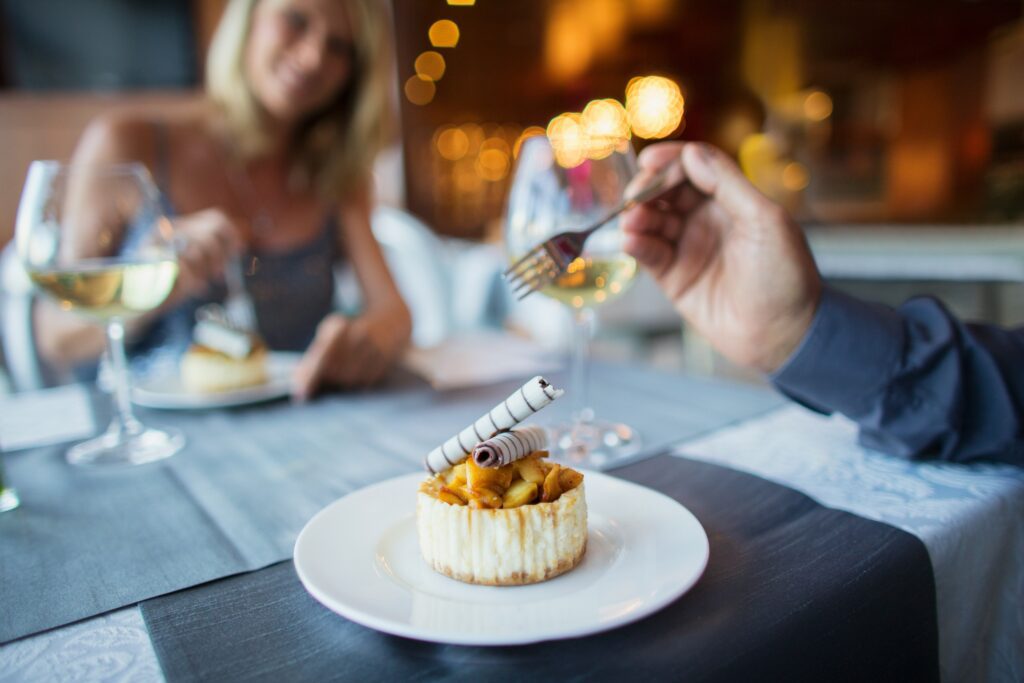 A close-up of a plated dessert with chocolate curls, with a blurred couple enjoying wine and dessert at a restaurant in the background.