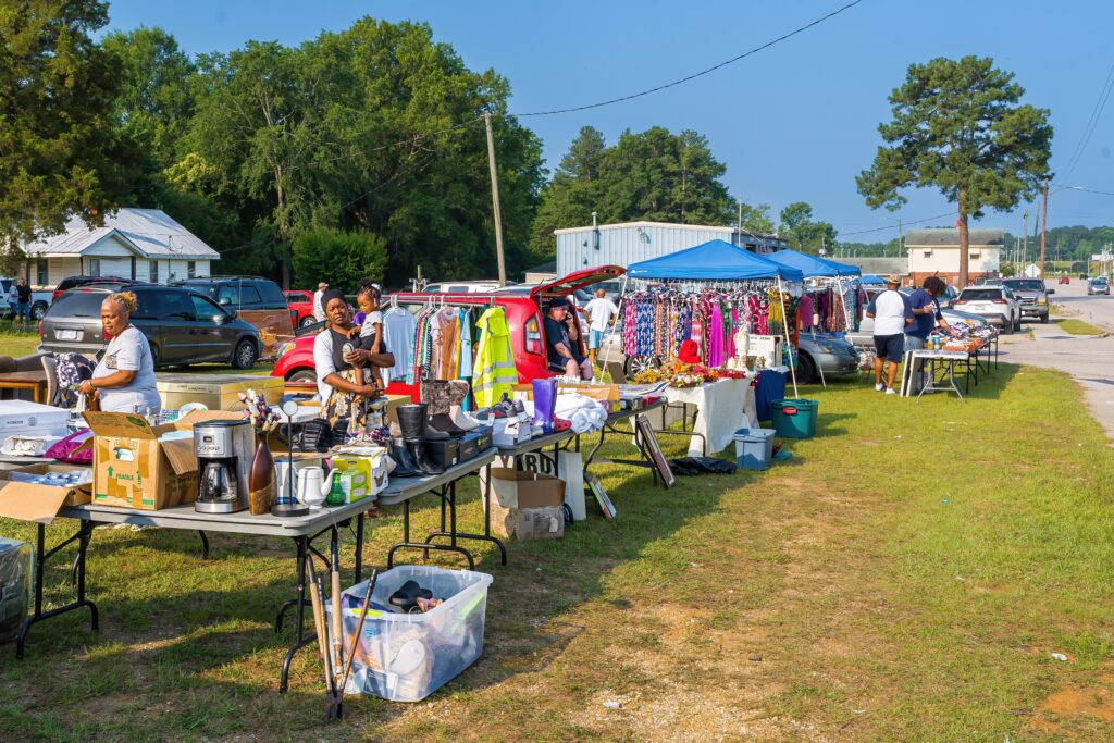 Outdoor flea market with tables displaying clothes, kitchen appliances, and various items. People are browsing and selling under blue tents on a sunny day with cars and trees in the background.