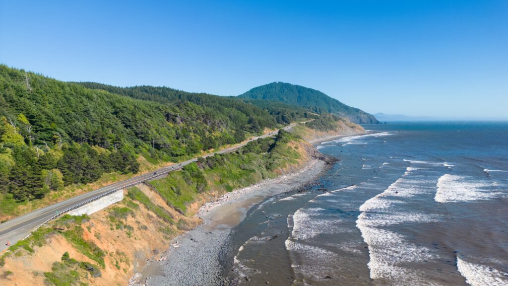 A scenic coastal highway runs alongside a rocky shoreline and forested hills under a clear blue sky, with gentle waves rolling onto the beach.