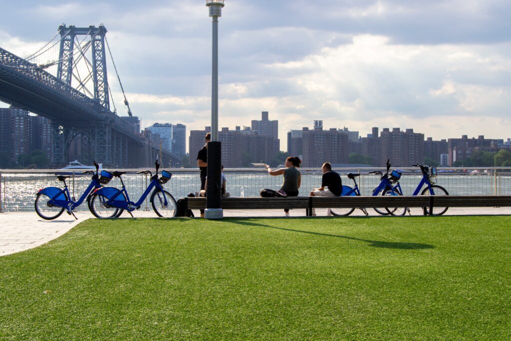 People sit and chat on benches by the water with blue rental bikes nearby, green grass in the foreground, and a large bridge and city buildings in the background under a partly cloudy sky.