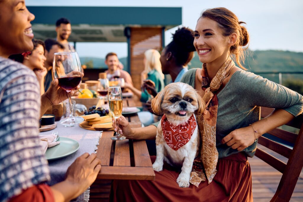 A smiling woman sits at an outdoor table with friends, holding a glass of wine. A small dog wearing a red bandana sits on her lap. The group appears to be enjoying a meal together.