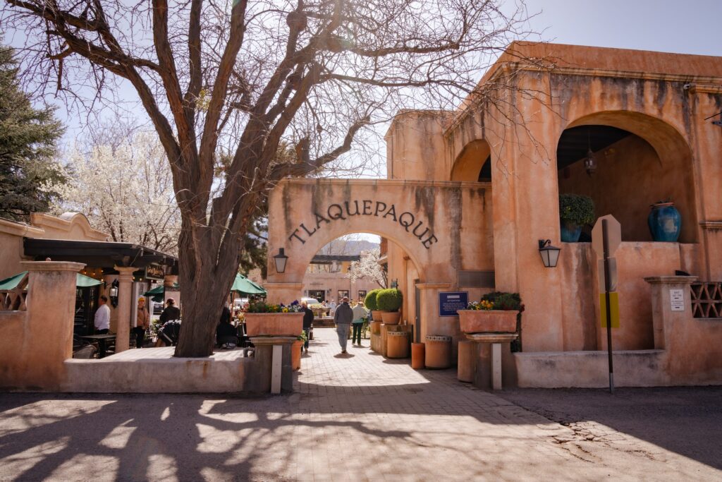 Entrance to Tlaquepaque, an adobe-style shopping and arts village, with people walking under the arched sign, potted plants, bare tree branches, and outdoor seating on a sunny day.